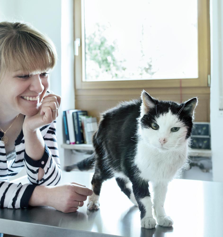 Eine lächelnde Frau mit blonden Haaren sitzt neben einer schwarz-weißen Seniorkatze, die in einer Tierheilpraktiker Praxis auf einem Tisch in einem hellen steht und Fürsorge und Aufmerksamkeit für die Tiergesundheit ausstrahlt.