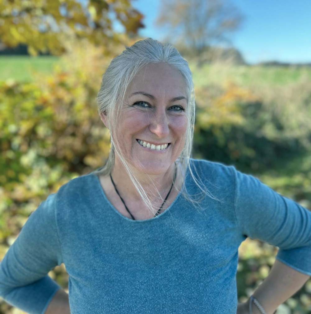 Eine lächelnde Frau mit langen grauen Haaren in einem blauen Shirt steht an einem sonnigen Tag im Freien.