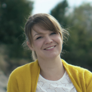 Franzisca mit hellbraunen Haaren und Pony lächelt im Freien. Sie trägt ein weißes Oberteil und einen senfgelben Pullover. Im Hintergrund sind verschwommen Bäume zu sehen.