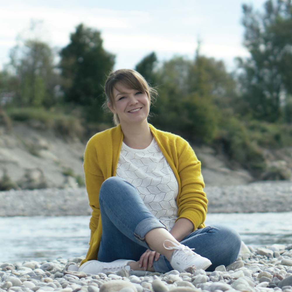 Franzisca mit Jeans und einer weißen Bluse mit gelber Strickjacke darüber, sitzt mit gekreuzten Beinen auf Kieselsteinen am Flussufer, lächelt in die Kamera. Im Hintergrund sind Bäume und ein bewölkter Himmel zu sehen, die eine friedliche Atmosphäre schaffen, die ihre Leidenschaft für die Tiergesundheit widerspiegelt.