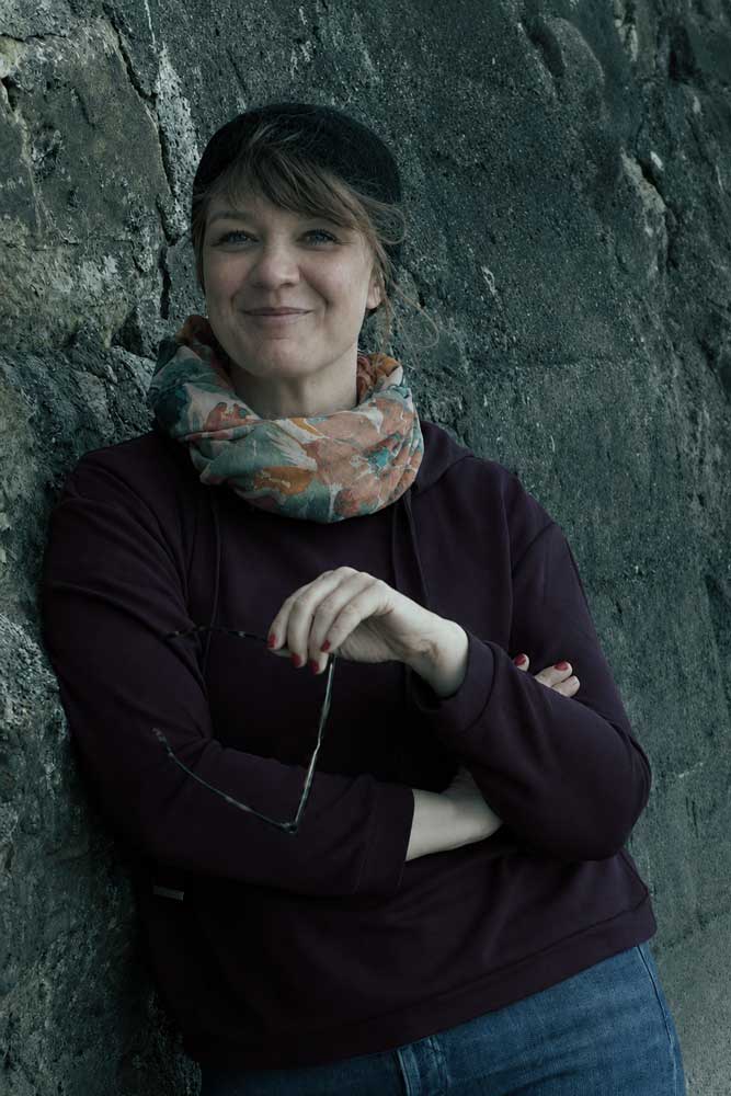 Franzisca mit einem geblümten Schal und einem dunklen Pullover steht vor einer Steinmauer, lächelt mit verschränkten Armen und hält eine Sonnenbrille in der Hand – ihre herzliche Ausstrahlung spiegelt ihr Engagement für die Tiergesundheit wider.