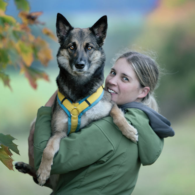 Eine blonde Frau in einer grünen Jacke lächelt, während sie draußen einen großen Hund im Arm hält, was deutlich ihre Freude an Tiergesundheit unterstreicht. Der Hintergrund ist verschwommen und mit grünen und herbstfarbenen Blättern übersät.