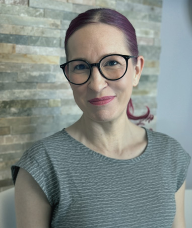 Eine Frau mit lila-pinken Haaren, die zu einem Pferdeschwanz zusammengebunden sind, mit einer runden schwarzen Brille und einem grau gemusterten Oberteil, die vor einer Wand aus Steinfliesen lächelt.