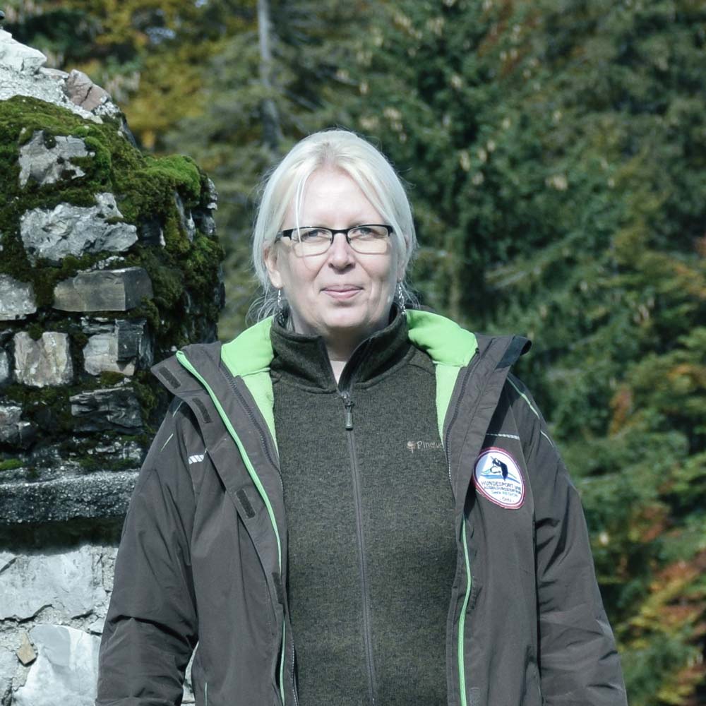 Eine Frau mit hellem Haar und Brille, die eine dunkle Jacke trägt, steht im Freien, im Hintergrund sind Bäume zu sehen.